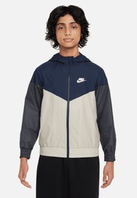 Jachetă Nike de tip windbreaker, cu glugă, design în blocuri de culori în nuanțe de bleumarin, gri și bej, cu manșoane elastice și fermoar frontal.