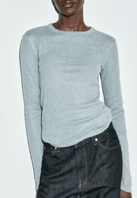 Camicia a maniche lunghe grigia chiara con scollatura rotonda, realizzata in tessuto morbido, abbinata a jeans di denim scuro con cuciture e tasche visibili.