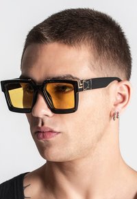 Occhiali da sole neri a forma quadrata con lenti gialle, caratterizzati da montature spesse e un design geometrico. Dettagli in metallo vicino alle tempie migliorano la struttura.