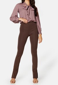 Fioletowa bluzka z dużą kokardą przy dekolcie i długimi rękawami, w połączeniu z brązowymi spodniami z wysokim stanem i bocznymi rozcięciami.