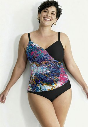 Lächelnde Frau mit kurzen lockigen Haaren, die Creolen und einen schwarzen Badeanzug mit einem bunten abstrakten Muster am Oberteil vor weißem Hintergrund trägt.