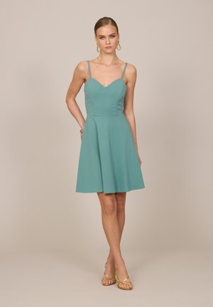 Femme debout portant une robe turquoise sans manches en ligne A avec de fines bretelles, assortie à des boucles d’oreilles créoles dorées et des talons dorés à lanières.