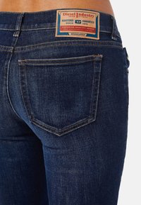Los jeans de mezclilla azul oscuro presentan un parche de marca en la cintura, costuras en contraste y dos bolsillos traseros con solapas puntiagudas.