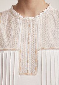 Blusa blanca con bordado texturizado y diseño plisado, con escote en forma de ondas y sutiles acentos beige.