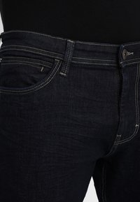 Dark blue denim jeans featuring a standard fit, contrasting yellow stitching, front pockets, and a button closure at the waist.