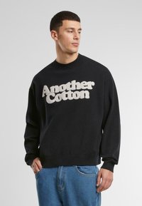 Sudadera negra con el texto "Another Cotton" en tamaño grande, en color blanco roto. Cuello redondo acanalado, mangas largas y corte relajado. Tejido de algodón suave.