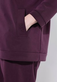 Sudadera de color púrpura oscuro con un bolsillo frontal, que presenta una textura suave y un discreto logo en strass en la manga.
