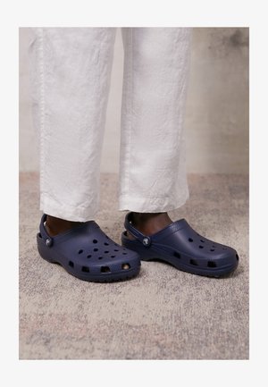 Crocs bleu marine en matériau flexible ; dotés de trous de ventilation et d'une sangle à l'arrière. Portés avec un pantalon blanc légèrement froissé.