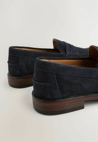 Mocasines de ante azul marino con una textura suave, tacón bajo y acentos de costura en la parte superior, con una suela de aspecto madera marrón.