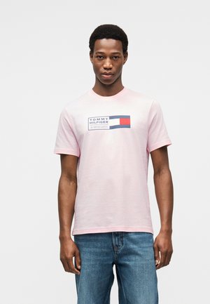 Νέος άνδρας που φοράει ένα ανοιχτό ροζ T-shirt Tommy Hilfiger με τζιν, στέκεται μπροστά από μια απλή ανοιχτό γκρι φόντο.