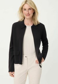 Schwarze Strickjacke mit Kragen, horizontalem Reißverschluss und Vordertasche. Über einem hellen Oberteil getragen und mit beigen Hosen kombiniert.