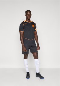 Sort fotballdrakt med oransje detaljer, kortermet og med logo. Sorte shorts og hvite sokker. Sorte fotballsko fullfører antrekket.