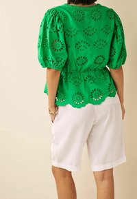 Top verde ricamato con motivi floreali a taglio, maniche a sbuffo e orlo scallop, abbinato a pantaloni corti in lino bianchi.