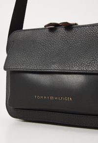 Geantă crossbody din piele neagră, cu un capac texturat, logo-ul auriu "TOMMY HILFIGER", secțiune inferioară netedă și un detaliu elegant al închiderii.