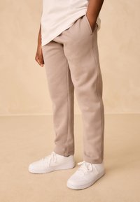 Pantalon de survêtement beige avec taille élastique et poches latérales, associé à des baskets blanches, vu de la taille vers le bas sur un fond neutre.