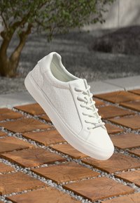 White sneaker with textured pattern on the side, round toe, and lace-up design. Smooth rubber sole and light cushioning visible.