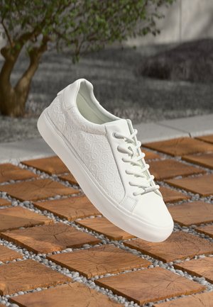 White sneaker with textured pattern on the side, round toe, and lace-up design. Smooth rubber sole and light cushioning visible.
