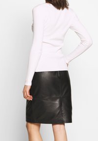 Mujer vestida con un suéter blanco ajustado de manga larga y una falda de cuero negra hasta la rodilla, de pie con una mano en la cadera.