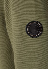 Olivgrön strukturerad tyg med vertikal ribbning, som har en rund svart patch med logotydelse på sidan.