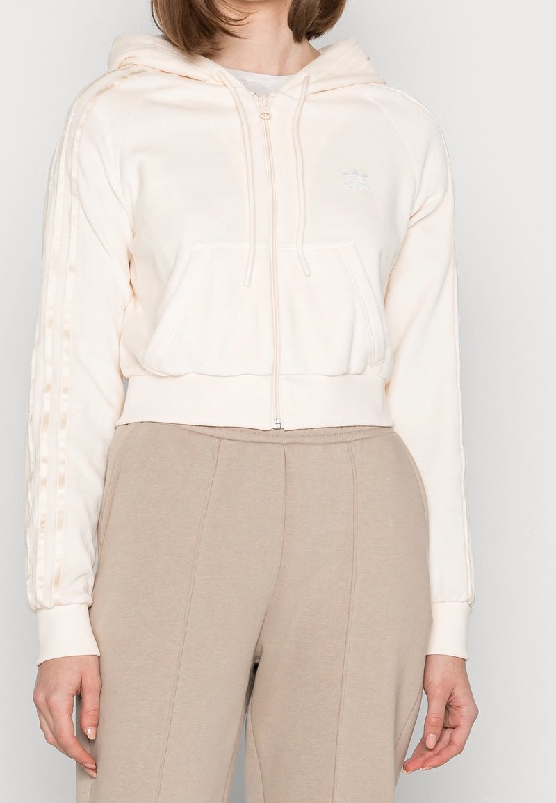 Kvinna i krämfärgad Adidas zip-up hoodie med ränder på ärmarna och beige höga byxor, med avslappnade händer vid sidorna.