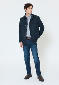 Young man standing, wearing a navy jacket, light blue button-up shirt, blue jeans, brown belt, and dark brown boots, against a plain white background.