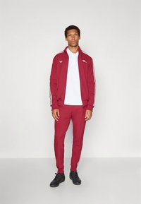 Fato de treino atlético vermelho com listras brancas, composto por uma jaqueta com fecho de correr e calças combinando. A jaqueta tem gola branca e detalhe de logótipo.