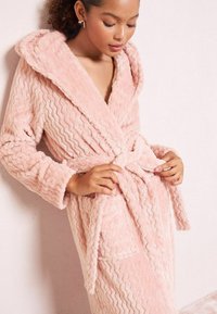 Jemný ružový fleecový župan s vlnitou textúrou, veľkou kapucňou a zodpovedajúcim opaskom. Obsahuje dlhé rukávy a bočné vrecká pre praktickosť.