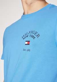 Detailní záběr osoby v modrém tričku se nápisem "HILFIGER NEW YORK" a červeno-bílo-modrým logem potištěným na hrudi.