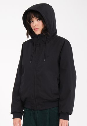 Jeune personne portant une veste noire à capuche avec fermeture éclair, debout devant un fond blanc uni, regardant légèrement sur le côté.
