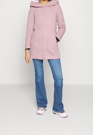 Person iført en lyserød hættetrøje, blå vide jeans og hvide sneakers med lyserøde detaljer, stående med den ene hånd i lommen.