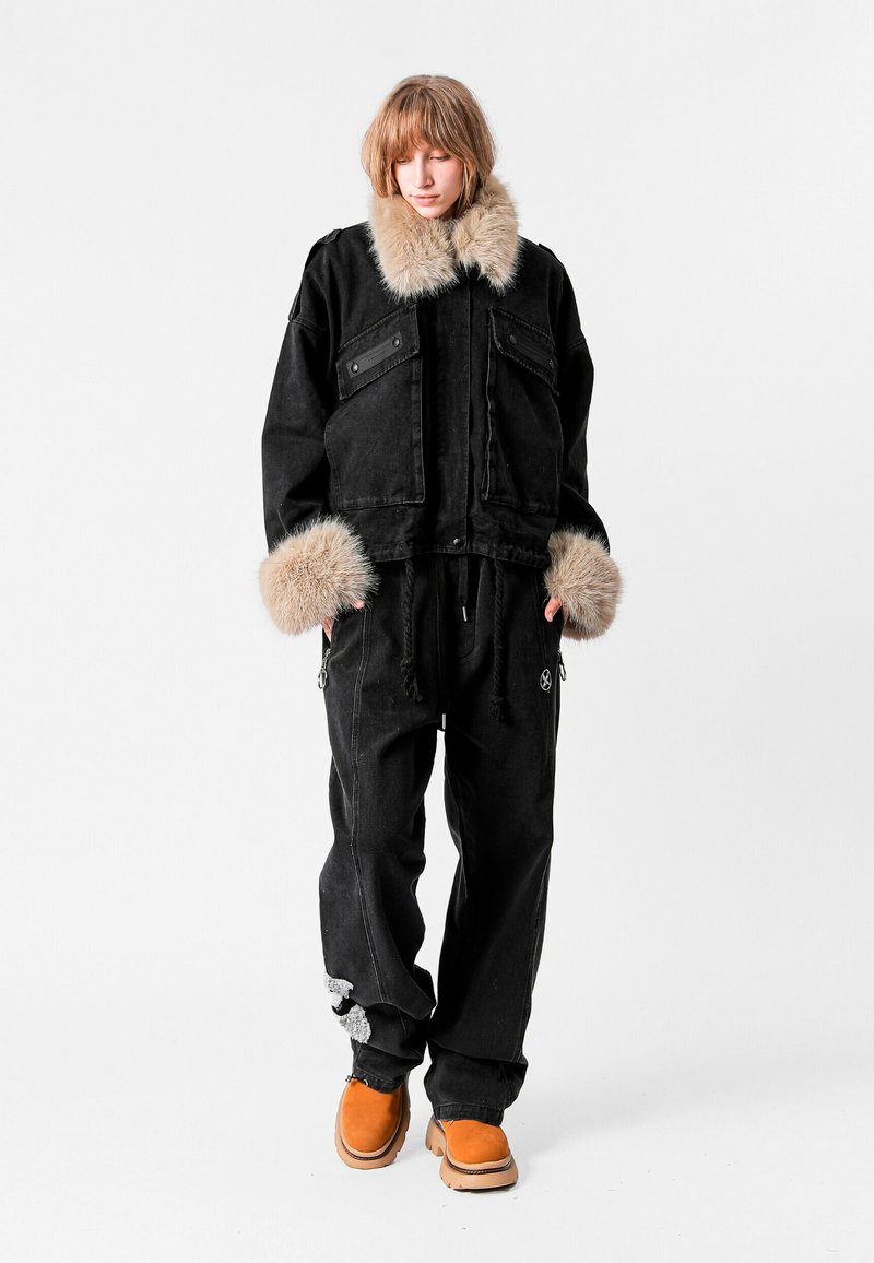 Chaqueta de mezclilla negra con cuello y puños de piel; combinada con pantalones negros holgados y calzado naranja. Presenta bolsillos con cremallera y detalles en contraste.
