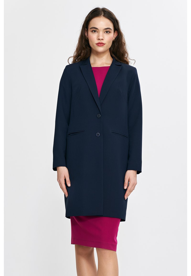 Granatowy długi płaszcz z dwoma guzikami, wyprofilowanymi klapami i bocznymi kieszeniami, zestawiony z dopasowaną, magentową sukienką pod spodem. Gładka tekstura tkaniny.