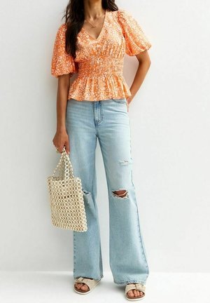 Vrouw die een oranje bloes met bloemmotief en pofmouwen draagt, gescheurde lichtblauwe spijkerbroek, beige gevlochten sandalen en een gevlochten crèmekleurige handtas vasthoudt.