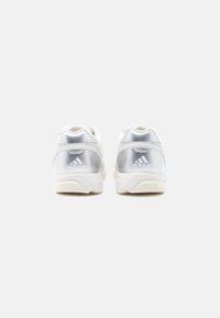 Bijele sportske cipele s srebrnim reflektirajućim dizajnom na peti i svijetlo obojenim potplatom, s naglascima logotipa sa strane.