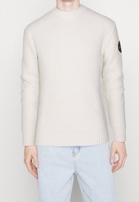 Vaaleanbeige ribbineuloksinen neulepusero pyöreällä pääntiellä ja pitkillä hihoilla. Vasemmassa hihassa on logotäplä. Valmistettu teksturoidusta kankaasta.