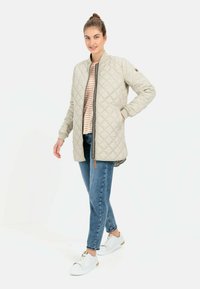 Hellbeige gesteppte Jacke mit Reißverschluss, Stehkragen und Seitentaschen, kombiniert mit einem gestreiften Hemd und blauen Jeans, getragen mit weißen Sneakers.