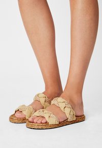 Vävda beigefärgade sandaler med flätade remmar, platta sisalsulor och texturerade detaljer. Öppen tå-design som framhäver den naturliga fotens form.