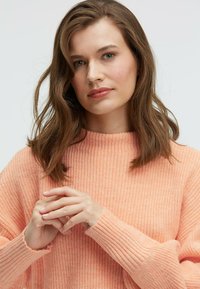 Textured, light peach sweater with a high neck and ribbed cuffs. The knit pattern features a soft, cozy finish. Hands clasped in front.
