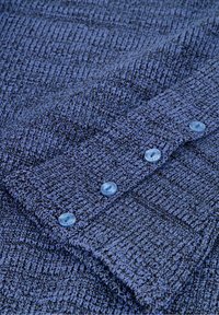 Blauw gebreide stof met een gestructureerd patroon. Bevat lichtblauwe knopen langs de manchetten. Het materiaal is zacht en flexibel.