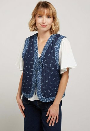 Femme aux cheveux blonds courts porte un chemisier blanc à manches courtes sous un gilet bleu marine à motifs floraux, debout, les mains près des hanches.