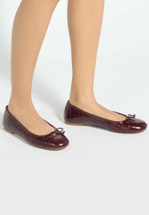 Jambes portant des ballerines rouge foncé embossées crocodile avec de petits nœuds décoratifs sur un fond clair.