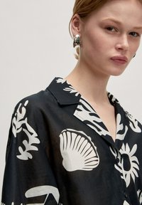 Schwarzes Hemd mit weißen Muschel- und Blumenmustern; verfügt über einen Reverskragen und Knöpfe; aus leichtem, fließendem Material gefertigt.