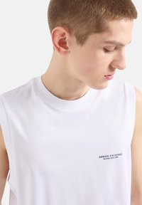 Biała koszulka na ramiączkach z bawełny, z okrągłym dekoltem i małym czarnym napisem logo na piersi. Gładka tekstura i minimalistyczny design.