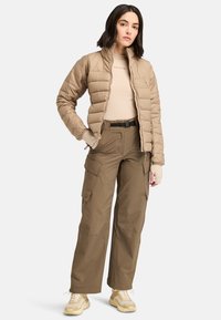 Giacca beige isolata con motivo trapuntato, abbinata a pantaloni cargo khaki con molte tasche e sneakers beige.