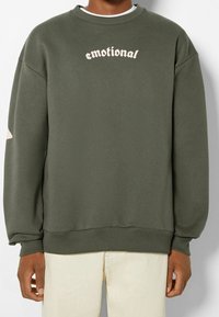 Grüner Pullover aus weichem Stoff, mit Rundhalsausschnitt, langen Ärmeln, gerippten Bündchen und einem „emotionalen“ Textdesign in Hellrosa.