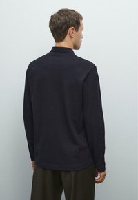 Mann, der von hinten mit einem schwarzen Langarmshirt und dunkler Hose vor einem einfarbigen hellgrauen Hintergrund steht.