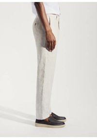Pantaloni in lino beige chiaro con taglio dritto, caratterizzati da una piega frontale, dettagli delle tasche e abbinati a scarpe nere slip-on.