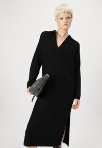 Musthave mustriba mustkootud kleit V-kaelusega, pikkade varrukatega ja külje lõhiku ning moodsa halli kotiga, mis on kinnitatud rihmaga.