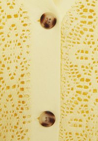 Gul kardígan med en stickad spetsdesign på ena sidan och en solid tyg på den andra sidan, med två runda, marmorliknande knappar för stängning.