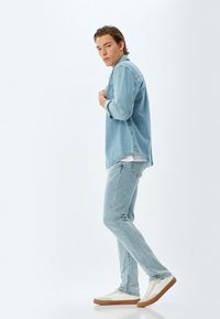 Camisa de denim azul claro sobre una camiseta blanca, combinada con jeans azul claro y zapatillas blancas con suelas marrones, ajuste estrecho, estilo casual.
